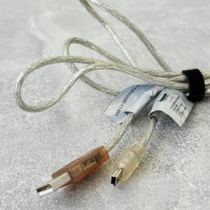 Puede incluir: Un cable USB 2.0 transparente y translúcido con un conector rectangular de tono marrón y un conector mini-USB más pequeño y claro. El cable está sujeto con una cinta de velcro negra e incluye una etiqueta blanca con USB 2.0 CERTIFIED.