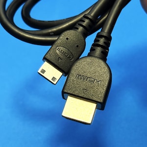 Może przedstawiać: Czarny kabel HDMI z dwoma różnymi złączami. Jedno złącze to standardowa wtyczka HDMI pełnej wielkości, a drugie to mniejsza wtyczka mini-HDMI. Obie czarne plastikowe obudowy złączy mają wytłoczone logo HDMI oraz małą strzałkę wskazującą kierunek.