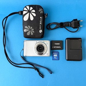 May include: Silver Canon digital camera with a black lens, a black camera case with white flower designs, a Canon battery, a charger, and an SD card. The camera has a 4x zoom and 12.1 megapixels.