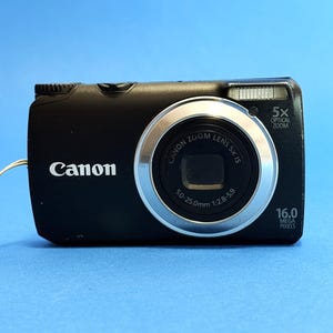 May include: A black Canon digital camera with a silver lens ring. The camera has a 5x optical zoom and 16.0 megapixels. The lens is a 50-25.0mm 1:2.8-5.9. The word "Canon" is printed in white on the front of the camera.