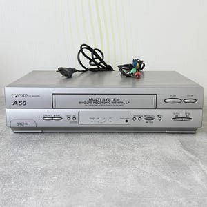 May include: A silver Sharp A50 multi-system VHS player with a front-loading cassette compartment, various control buttons, and indicator lights. The device features text including Multi System, 8 Hours Recording with PAL LP, and VHS HQ, accompanied by a black power cord and a set of multi-colored RCA cables.