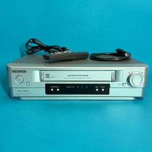 Könnte beinhalten: Silberner Samsung VHS-Player mit Fernbedienung und schwarzen Kabeln. Der Player hat einen VHS-Kassetten-Einschub und Bedientasten. Der Text "x400 SUPER SPEED REWIND" ist auf der Vorderseite sichtbar.