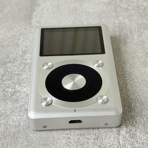 May include: A rectangular silver digital music player featuring a black circular navigation wheel with a central silver button. The device includes a dark screen at the top, four small silver control buttons surrounding the wheel, and a charging port.