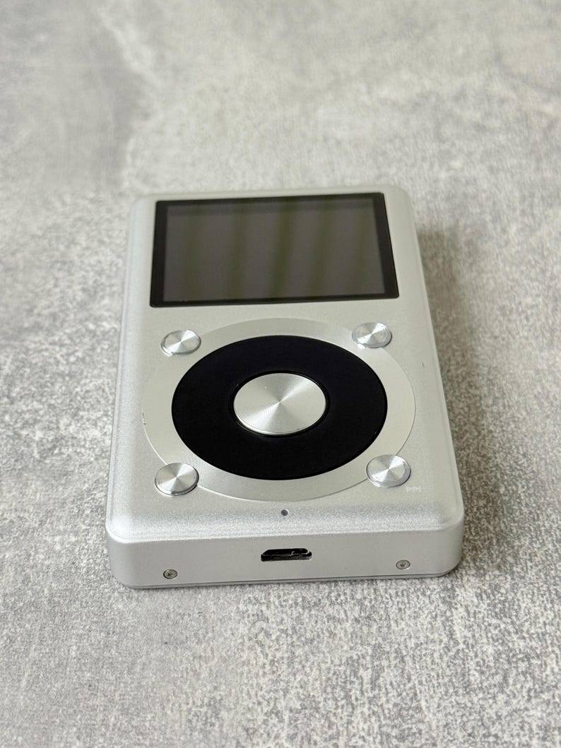 May include: A rectangular silver digital music player featuring a black circular navigation wheel with a central silver button. The device includes a dark screen at the top, four small silver control buttons surrounding the wheel, and a charging port.