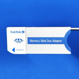 Könnte beinhalten: Ein silberner und weißer Memory Stick Duo Adapter mit dem Sandisk-Logo und dem Wort "INSERT" aufgedruckt. Der Adapter hat ein blaues Etikett mit der Aufschrift "Memory Stick Duo Adaptor".