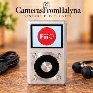 Puede incluir: Un reproductor de música digital plateado con una rueda de control circular negra y una pantalla que muestra un círculo rojo con el texto blanco FiiO. El dispositivo viene acompañado de un cable de carga USB negro y auriculares negros.