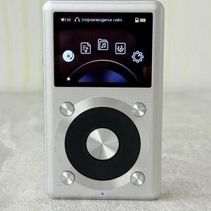 May include: A silver digital music player with a rectangular screen displaying icons and a black circular navigation wheel. The device features four small silver buttons surrounding the wheel and a small blue indicator light at the bottom centre.