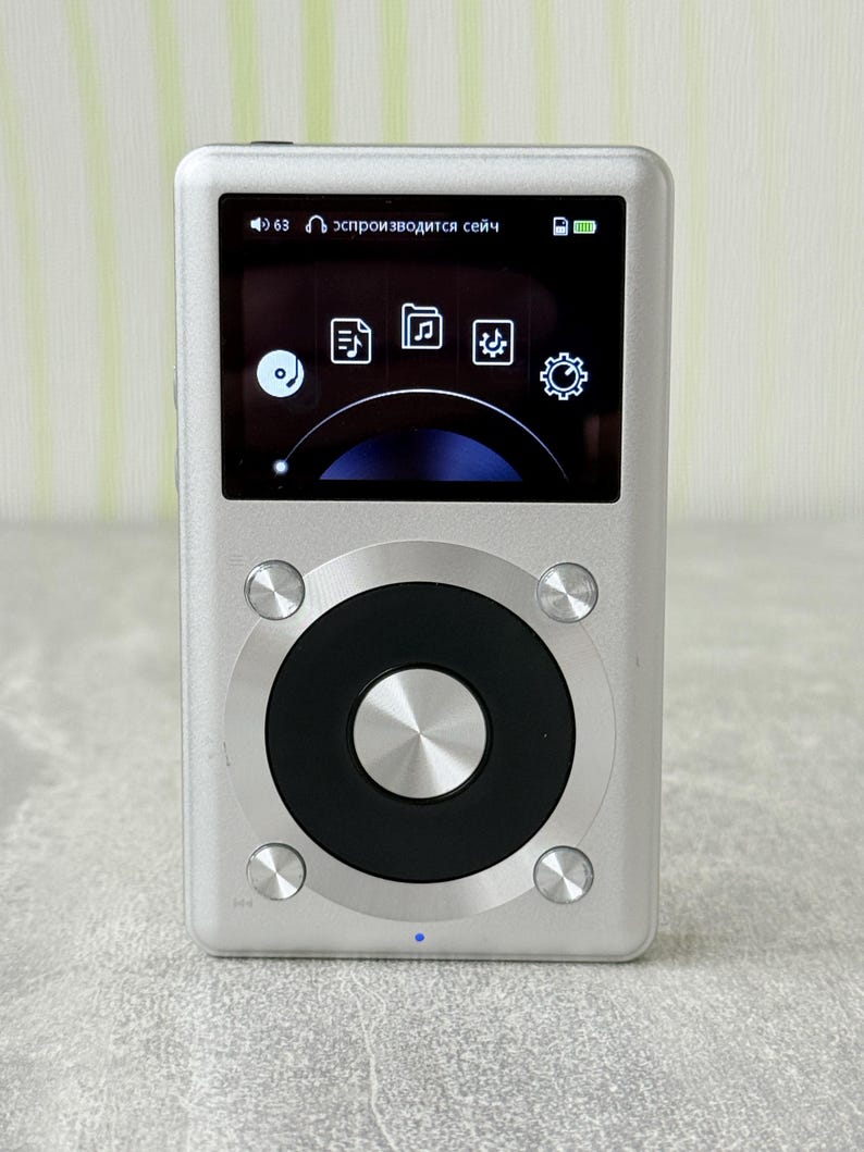 May include: A silver digital music player with a rectangular screen displaying icons and a black circular navigation wheel. The device features four small silver buttons surrounding the wheel and a small blue indicator light at the bottom centre.