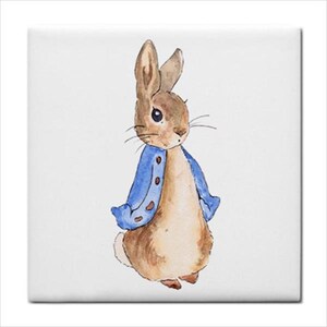 May include: A square ceramic tile featuring a watercolor illustration of a brown rabbit standing upright. The rabbit is wearing a blue jacket with visible brown buttons. The illustration is centered on a plain white background with clean edges.
