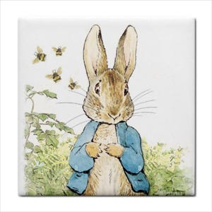 May include: A square decorative tile featuring a watercolor illustration of a brown rabbit wearing a blue jacket. The rabbit stands in a patch of green foliage and yellow flowers, with four small bees flying in the upper left corner of the scene.