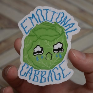 Puede incluir: Una pegatina blanca con una col verde con una cara triste y ojos azules en forma de lágrima. El texto "Emotional Cabbage" está escrito en azul sobre la col y "Cabbage" está escrito en azul debajo de la col.