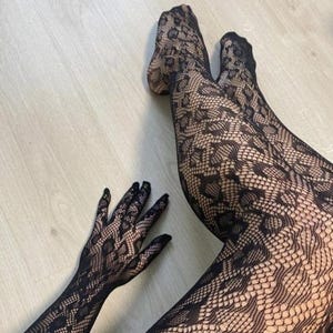 Könnte beinhalten: Schwarze Spitzenstrumpfhose und ein passender Handschuh werden vor einem hellen Holzmaserungshintergrund gezeigt. Das komplizierte Muster der Spitze erzeugt einen strukturierten Effekt und verleiht dem Gesamtbild einen Hauch von Eleganz und Raffinesse.