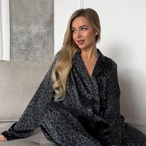 Puede incluir: Un conjunto de pijama gris oscuro con estampado de leopardo. El conjunto incluye una camisa con botones y pantalones a juego. La persona lleva zapatillas negras y peludas. El pijama está hecho de un material suave y sedoso.