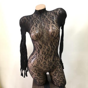 Puede incluir: Body de encaje negro con cuello alto y mangas largas. El body presenta un estampado de piel de serpiente y se exhibe en un maniqu&iacute;. La prenda est&aacute; dise&ntilde;ada para ser ajustada y est&aacute; hecha de un material transparente.