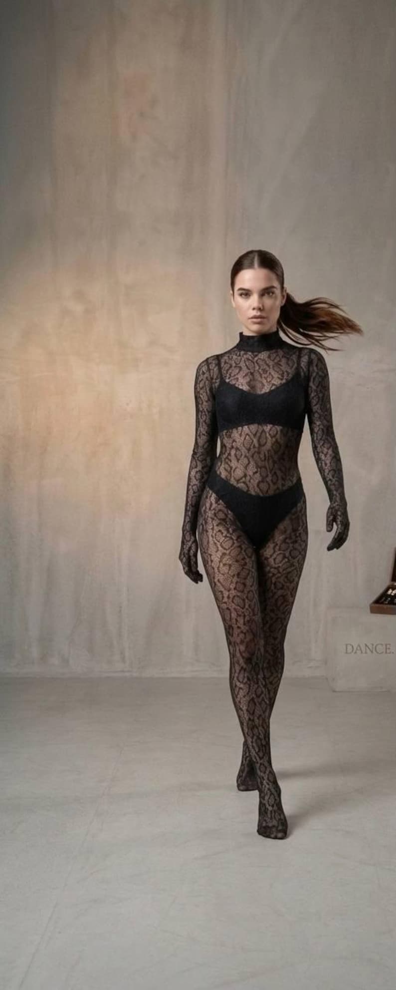 Puede incluir: Un body negro de cuello alto con estampado de piel de serpiente, combinado con sujetador, ropa interior y guantes a juego. La modelo camina. La palabra "DANCE" es visible en el fondo.