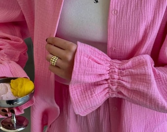 May include: A bright pink long-sleeved button-down shirt with a textured fabric and gathered, ruffled cuffs, worn over a white ribbed tank top featuring a small black logo on the chest. A gold ring with three stacked spheres is worn on a finger.