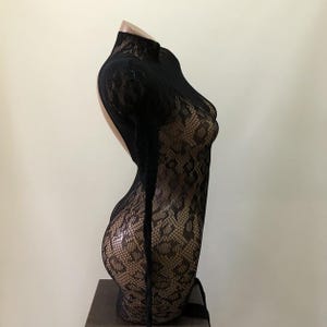 Puede incluir: Body de encaje negro con cuello alto y dise&ntilde;o de espalda abierta. El body se exhibe en un maniqu&iacute;, mostrando el intrincado patr&oacute;n floral del encaje. La prenda tiene mangas largas y una silueta ajustada.