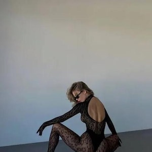 Puede incluir: Body de encaje negro con mangas largas y espalda abierta. El conjunto incluye medias y guantes estampados a juego. La modelo lleva tacones altos negros y gafas de sol. El look general es una declaraci&oacute;n de moda elegante y atrevida.