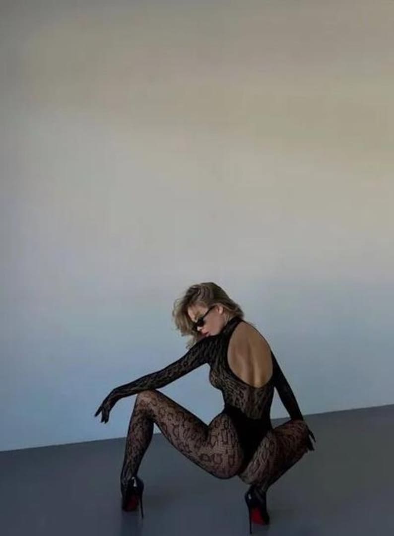 Puede incluir: Body de encaje negro con mangas largas y espalda abierta. El conjunto incluye medias y guantes estampados a juego. La modelo lleva tacones altos negros y gafas de sol. El look general es una declaraci&oacute;n de moda elegante y atrevida.