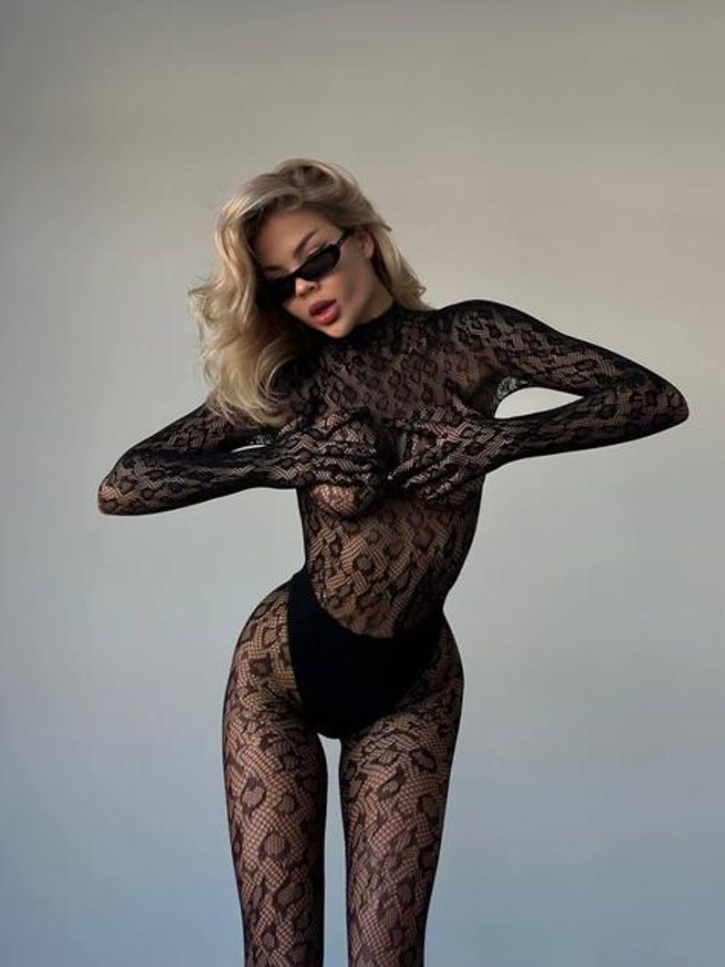 Puede incluir: Un body de encaje negro con mangas largas y cuello alto. El body presenta un dise&ntilde;o estampado y se combina con una parte inferior negra. La modelo lleva gafas de sol negras.