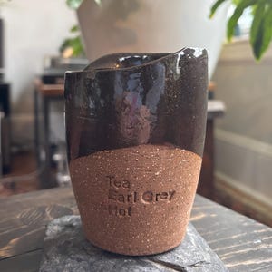 Puede incluir: Taza de cerámica hecha a mano con la parte superior esmaltada en marrón oscuro y una base marrón sin esmaltar y texturizada. Las palabras "Tea Earl Grey Hot" están impresas en la base. La taza está sobre un posavasos de piedra gris.