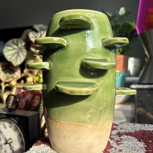 May include: A cylindrical ceramic vase with a glossy green glaze on the upper portion and a matte beige base. The exterior features several protruding, shelf-like horizontal ceramic discs extending from the sides of the vessel in a staggered pattern.