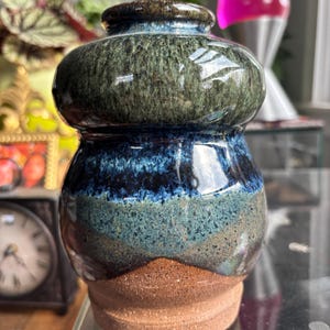 May include: A ceramic vase with a bulbous top and a rounded body, featuring a glossy glaze in shades of forest green, deep blue, and teal. The base is made of unglazed, textured terracotta-colored clay, contrasting with the smooth, shiny upper section.