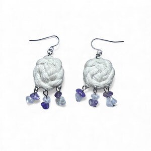 May include: A pair of handmade earrings featuring a woven white design with dangling purple and blue gemstones. The earrings have silver-colored hooks for pierced ears. The gemstones are various shapes and sizes.