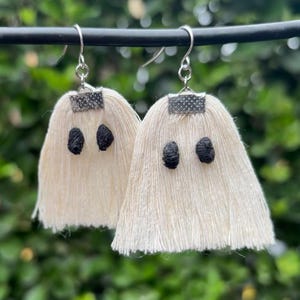May include: A pair of handmade ghost earrings. Each earring features a cream-colored, fringed body with two black, embroidered eyes. The earrings hang from silver-colored hooks and are perfect for Halloween.