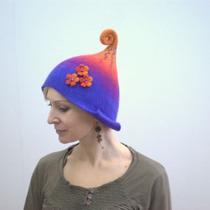 Peut inclure: Une femme portant un chapeau en feutre violet et orange avec un bord recourbé et des fleurs en feutre orange. Le chapeau est un design unique et fantaisiste.