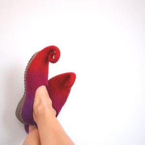 May include: A pair of red and purple felt slippers with pointed toes and curled up heels. The slippers are worn on a person's feet.