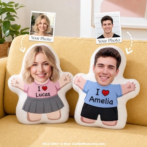 Könnte beinhalten: Zwei individuell geformte Plüschkissen mit aufgedruckten Gesichtern einer blonden und einer dunkelhaarigen Person. Die blonde Figur trägt ein rosa T-Shirt mit der Aufschrift 'I heart Lucas' und einen grauen Rock. Die dunkelhaarige Figur trägt ein blaues T-Shirt mit der Aufschrift 'I heart Amelia' und eine schwarze Shorts.