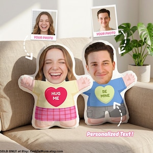 Puede incluir: Dos cojines de peluche con formas personalizadas que muestran los rostros impresos de una mujer y un hombre. El cojín de la mujer tiene una camiseta amarilla con un corazón rojo que dice HUG ME y una falda de cuadros rosa. El cojín del hombre tiene una camiseta azul con un corazón verde que dice BE MINE.