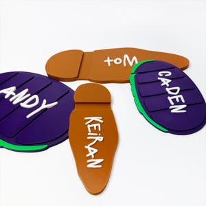 Può includere: Quattro etichette colorate con nomi di varie forme. Le etichette sono marroni, viola e verdi, con testo bianco che dice "TOM", "KEIRAN", "ANDY" e "CADEN". Le etichette sono disposte su uno sfondo bianco.