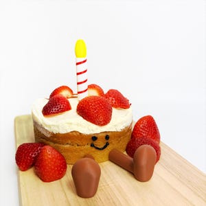 May include: A small round cake with white frosting, topped with fresh strawberries and a single candle. The cake has a smiling face drawn on it. Several whole strawberries and two brown objects are on a wooden board.