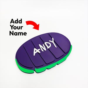 Op de afbeelding: Een paars en groen gepersonaliseerd naamlabel met de naam "ANDY" in witte letters. Het label heeft een gestructureerd oppervlak en een groene rand. De tekst "Voeg je naam toe" staat boven het label met een rode pijl die naar het label wijst.