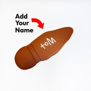 Puede incluir: Un objeto marrón con forma de zapato con el texto "toM" en blanco. Las palabras "Add Your Name" están encima de la forma del zapato con una flecha roja que apunta al zapato.