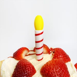 May include: A close-up of a cake with a yellow and white candle with red stripes. The cake is topped with white frosting and sliced strawberries. The background is plain white.
