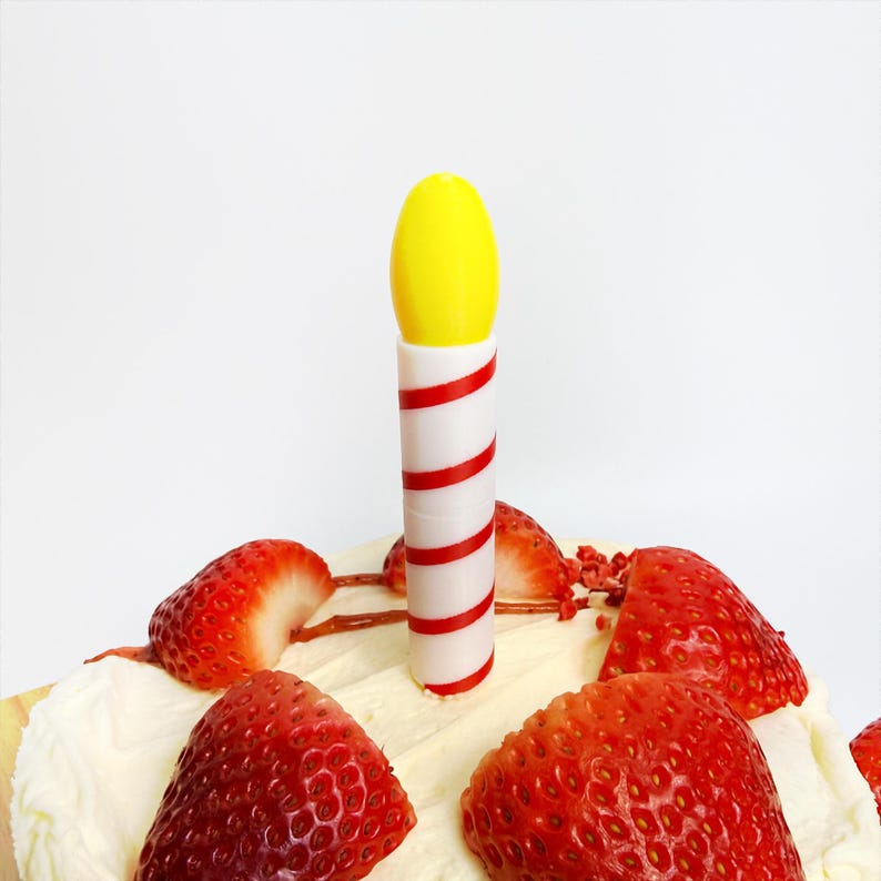 May include: A close-up of a cake with a yellow and white candle with red stripes. The cake is topped with white frosting and sliced strawberries. The background is plain white.