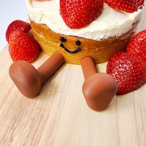 May include: A novelty cake decorated with a smiley face and brown legs, alongside fresh strawberries. The cake features a white frosting layer and is presented on a wooden board, creating a fun and playful scene.
