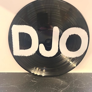 May include: A black vinyl record featuring the white letters "DJO". The record has grooves and is displayed against a light background. The record is a decorative item, possibly for wall art.