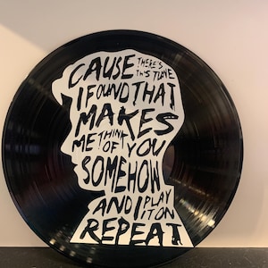 May include: A black vinyl record with a white silhouette of a person's head and shoulders. The record features the text "Cause I found that makes me think of you somehow and I play it on repeat."