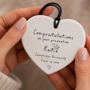 May include: A white heart-shaped ceramic ornament featuring black text that reads Congratulations on your graduation, Katie, Cambridge University, Class of 2026, with a small graduation cap icon and a heart symbol, suspended by a black cord.
