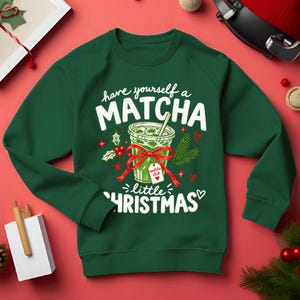 May include: A dark green long-sleeve sweatshirt featuring a graphic of a matcha drink in a clear cup with a red bow and a tag that reads "matcha club". The text "have yourself a MATCHA little CHRISTMAS" is printed in white around the illustration.