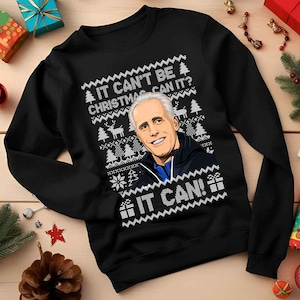 May include: Black sweatshirt with a festive design. The design includes the text "IT CAN'T BE CHRISTMAS CAN IT? IT CAN!" and a portrait of a man. The design also features Christmas trees, reindeer, and snowflakes.