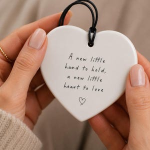 May include: A white heart-shaped ceramic ornament featuring a black string loop at the top. The surface is printed with the black text: A new little hand to hold, a new little heart to love, followed by a small hand-drawn heart symbol at the bottom.