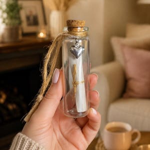 May include: A small clear glass bottle with a cork stopper, containing a rolled white paper scroll tied with twine. A silver-coloured metal heart charm hangs from the neck of the bottle, which is wrapped in a piece of natural jute twine for hanging.
