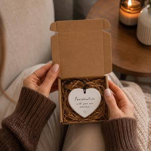 May include: A white ceramic heart-shaped ornament with a black string, nestled in a brown cardboard gift box filled with shredded paper. The ornament features the black printed text: Personalise with your own message, accompanied by a small heart icon.