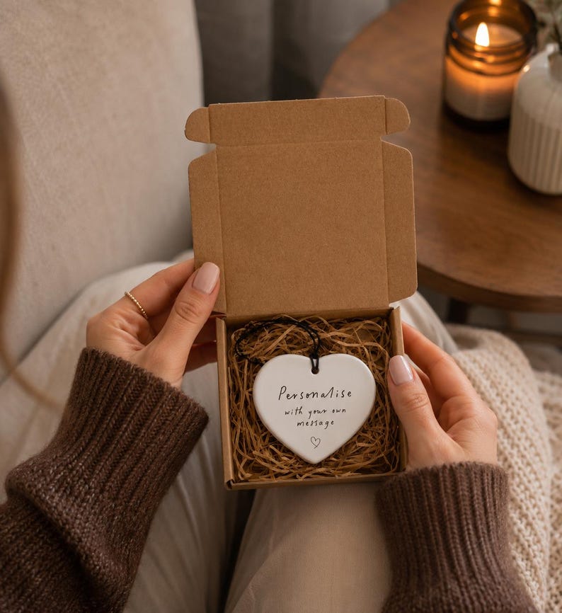 May include: A white ceramic heart-shaped ornament with a black string, nestled in a brown cardboard gift box filled with shredded paper. The ornament features the black printed text: Personalise with your own message, accompanied by a small heart icon.