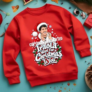 May include: A bright red long-sleeve sweatshirt featuring a graphic of a person wearing a Santa hat and a white-trimmed red coat. The text reads "Iraola Want For Christmas Is You" in white letters, surrounded by green holly leaves and white snowflakes.